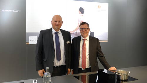 Zwei Geschäftsleute im Anzug stehen lächelnd an einem modernen Küchenmessestand mit Induktionskochfeld, Topf und Design‑Hinterwand mit Red-Dot-Award-Hinweis