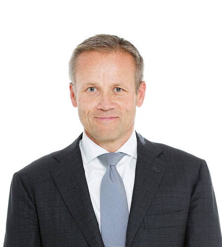 Mittelalterlicher Mann im dunklen Anzug mit hellblauer Krawatte lächelt dezent vor weißem Hintergrund