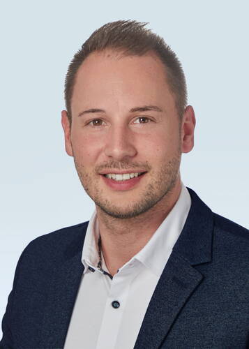 Junger Mann im Sakko und weißem Hemd lächelt freundlich in die Kamera vor hellem Hintergrund