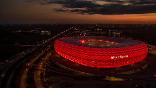 Leuchtend rot illuminierte Allianz Arena in München bei einsetzender Dämmerung mit vorbeifließendem Autobahnverkehr