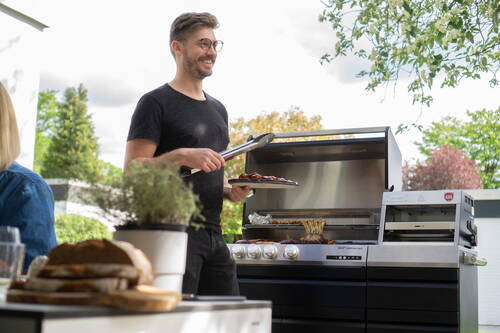 Mann mit Grillzange und Servierplatte steht lächelnd vor einem modernen Gasgrill im Garten und bereitet Fleisch für ein entspanntes Outdoor-Essen zu.