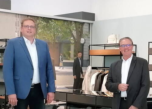 Zwei lächelnde Männer im Anzug stehen in einem modernen Bekleidungsgeschäft vor Kleiderständern und Regalen mit Jacken und Accessoires.