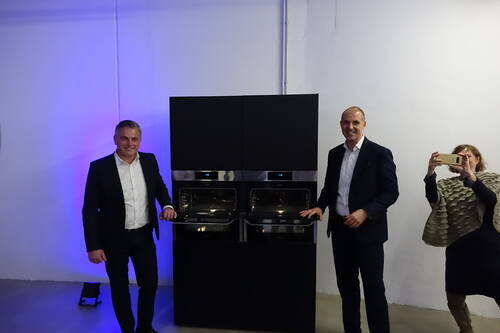 Zwei Männer in Anzügen präsentieren in einem Ausstellungsraum ein schwarzes Doppel-Backofensystem, während eine Frau im Hintergrund mit ihrem Smartphone ein Foto macht