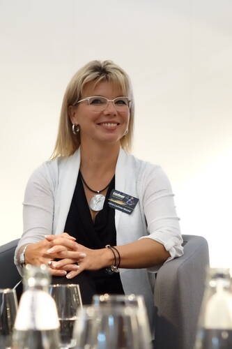 Lächelnde Frau mit Brille und Namensschild sitzt entspannt in einem grauen Sessel bei einer Veranstaltung im Konferenzraum.