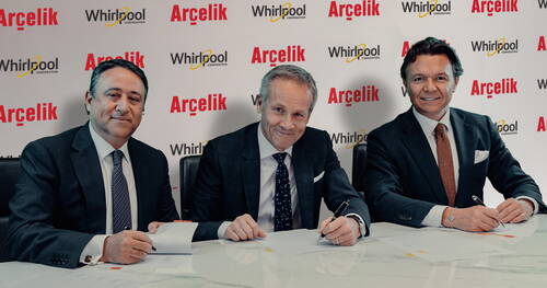 Drei Geschäftsleute in Anzügen sitzen an einem Konferenztisch und unterzeichnen lächelnd offizielle Dokumente vor einer Hintergrundwand mit den Logos von Arçelik und Whirlpool.