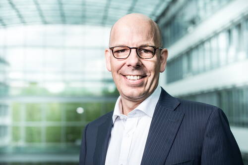 Lächelnder, glatzköpfiger Mann mit Brille im Business-Anzug steht in einer modernen, verglasten Büroumgebung