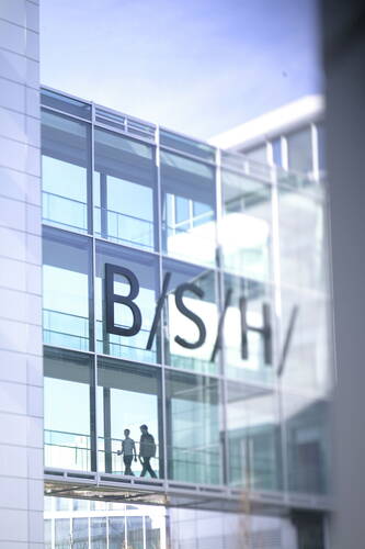 Zwei Personen gehen durch eine verglaste Bürobrücke im modernen BSH-Gebäude mit großer Firmenaufschrift an der Fassade