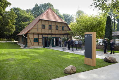 Historisches Fachwerkhaus mit rotem Ziegeldach, gepflegter Rasenfläche und elegant gekleideten Menschen bei einer Veranstaltung neben einer modernen Holzstele mit Firmenlogo.