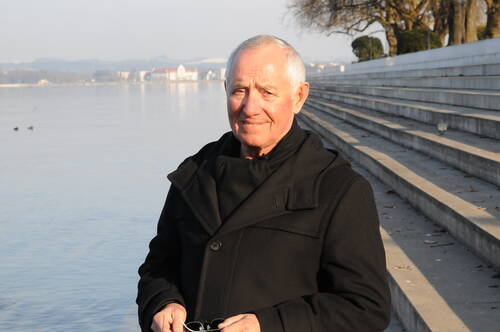 Älterer Mann in dunklem Mantel steht lächelnd an einem ruhigen Seeufer vor weitläufigen Betonstufen an einem klaren Wintertag.