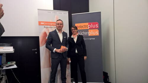 Zwei Geschäftsleute stehen lächelnd im Anzug zwischen EK/servicegroup- und electroplus küchenplus‑Rollups in einem Konferenzraum.