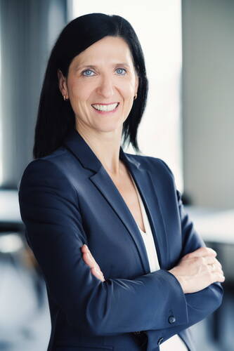 Selbstbewusste Geschäftsfrau mit dunklen Haaren und blauem Blazer steht mit verschränkten Armen lächelnd in einem modernen Büro.