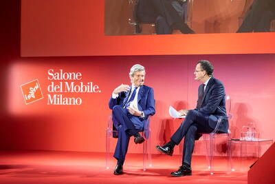 Zwei elegant gekleidete Männer diskutieren lächelnd auf einer roten Bühne beim Salone del Mobile in Mailand, sitzend auf transparenten Stühlen vor dem Eventlogo.