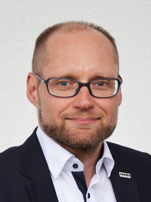 Lächelnder, glatzköpfiger Mann mit Brille und Bart in Anzug und weißem Hemd vor heller Wand