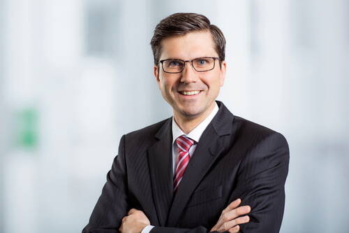Geschäftsmann mit Brille und Anzug verschränkt lächelnd die Arme vor einem hellen, unscharfen Bürohintergrund