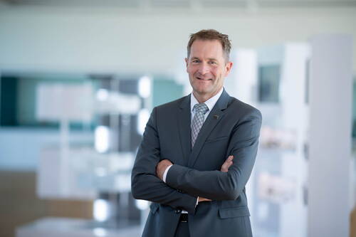 Geschäftsmann mittleren Alters im grauen Anzug mit verschränkten Armen lächelt selbstbewusst in einem modernen, hellen Büro.