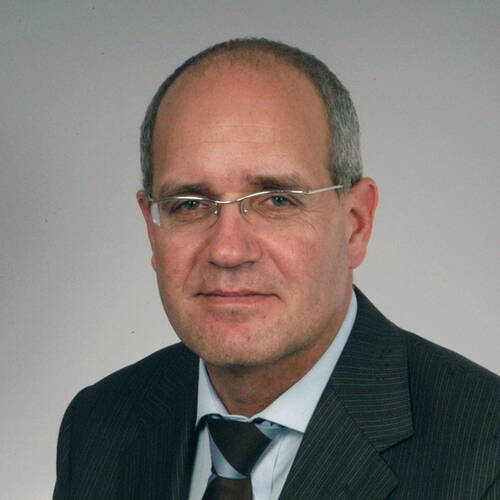 Älterer Mann mit Brille und Glatze in dunklem Anzug und Krawatte vor neutral grauem Hintergrund