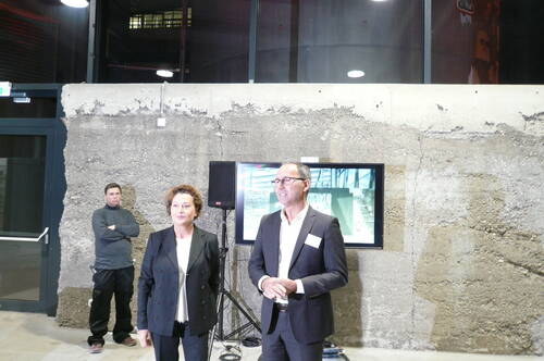 Zwei Geschäftsleute stehen vor einer rohen Betonwand und sprechen bei einer Präsentation, während im Hintergrund ein Mann in Arbeitskleidung vor einem Bildschirm mit Architekturbildern steht.