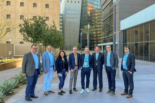 Gruppe von acht Geschäftsleuten steht lächelnd in einer modernen Hofpassage vor dem World Market Center mit hohen Glasfassaden und Bäumen im Hintergrund