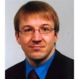 Mann mit kurzem, hellbraunem Haar, Brille und Anzug mit blauem Hemd blickt neutral in die Kamera vor hellem Hintergrund