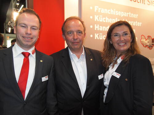 Drei lächelnde Geschäftsleute in Anzügen posieren auf einer Messe vor einem Stand mit der Aufschrift „Franchisepartner“ und „Küchenfachberater“.