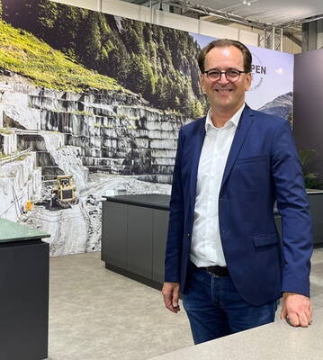 Geschäftsmann im blauen Sakko steht lächelnd vor einem Messestand mit großformatigem Steinbruch-Panoramabild im Hintergrund