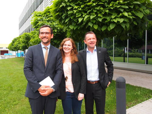 Drei lächelnde Berufstätige in Businesskleidung stehen nebeneinander vor einem modernen Bürogebäude mit begrünten Bäumen im Hintergrund