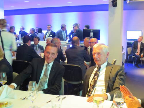 Elegante Abendveranstaltung mit festlich gekleideten Geschäftsleuten, die an rund gedeckten Tischen sitzen, sich unterhalten und Wein trinken, während im Hintergrund weitere Gäste stehen und diskutieren.