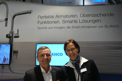 Zwei lächelnde Messemitarbeitende in Businesskleidung präsentieren vor einem BLANCO-Stand moderne Smart-Armaturen mit Werbeslogan im Hintergrund.