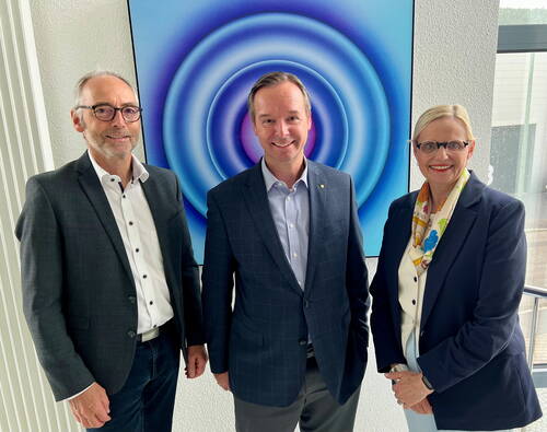 Drei geschäftlich gekleidete Personen stehen lächelnd nebeneinander in einem modernen Büro vor einem großen, blau-violetten Kunstwerk mit konzentrischen Kreisen.