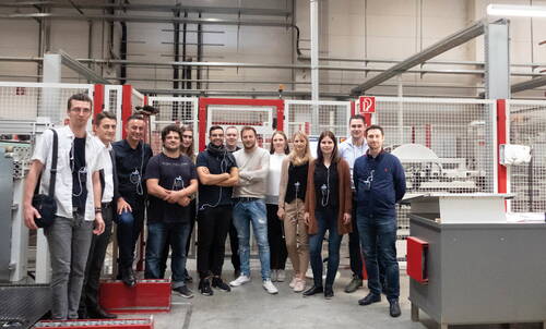 Gruppe von Mitarbeiterinnen und Mitarbeitern steht mit Besucher-Headsets in einer industriellen Produktionshalle vor einer Maschine und blickt lächelnd in die Kamera
