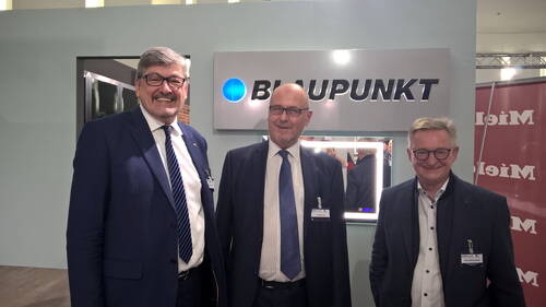 Drei lächelnde Männer im Anzug stehen nebeneinander vor einem Blaupunkt-Messestand in einer hell beleuchteten Ausstellungshalle.