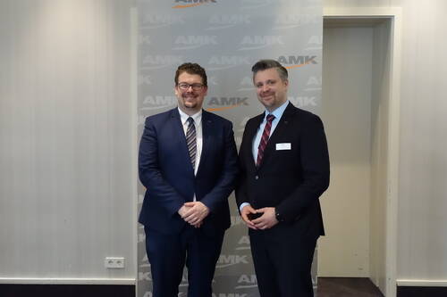 Zwei lächelnde Männer im Anzug posieren nebeneinander vor einer hellen Wand mit AMK‑Logo in einem Konferenzraum