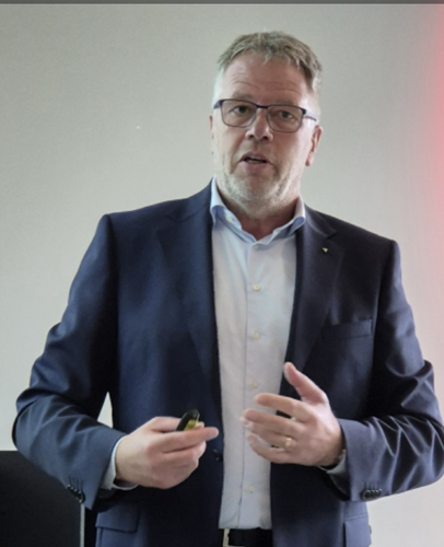 Mittelalterlicher Mann im Anzug hält einen Präsentationsklicker in der Hand und spricht gestikulierend vor neutralem Hintergrund