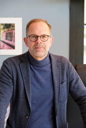 Mittelalterlicher Mann mit Brille und grauem Bart steht in dunkelblauem Rollkragenpullover und Sakko vor einer grauen Wand mit gerahmtem Bild im Hintergrund