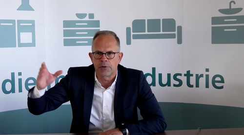 Mann im Anzug spricht gestikulierend vor einer Wand mit Piktogrammen von Haushaltsmöbeln und dem Schriftzug „Industrie“.