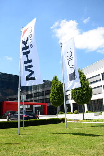 Zwei Fahnen mit den Schriftzügen „MHK Group“ und „unic.“ wehen vor einem modernen Bürogebäude mit Glasfassade, rotem Eingangsportal und gepflegtem Grünbereich unter blauem Himmel.