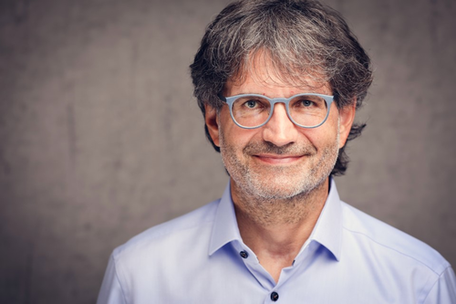 Mittelalterlicher Mann mit grauen Locken und hellblauer Brille lächelt freundlich in die Kamera vor neutralem Hintergrund.
