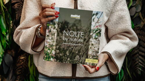 Person mit rotem Nagellack hält den „NOLTE by nature – Sustainability Report 2023“ vor einem grünen, pflanzlichen Hintergrund in die Kamera.