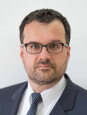 Mittelalterlicher Mann mit kurzer dunkler Frisur, Brille und Dreitagebart im Anzug und gestreifter Krawatte blickt ernst in die Kamera vor neutralem Hintergrund