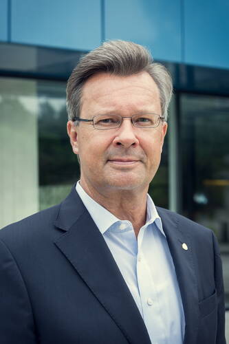 Älterer Mann mit Brille im dunklen Anzug und offenem Hemdkragen blickt ernst in die Kamera vor moderner Glasfassade