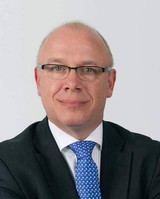 Glatzköpfiger Mann mit Brille im dunklen Anzug und blauer Krawatte blickt ernst in die Kamera vor neutralem Hintergrund.