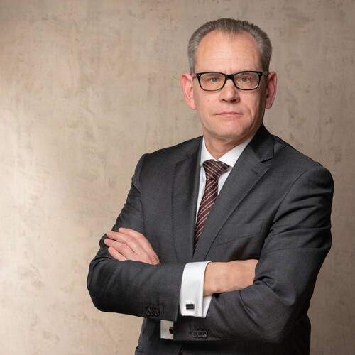 Ernst dreinblickender Geschäftsmann mittleren Alters im dunklen Anzug mit verschränkten Armen vor neutralem Hintergrund