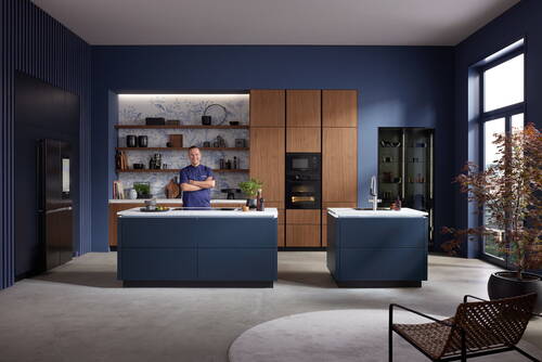 Mann in moderner, blau-goldener Designküche steht an Kochinsel mit Marmorplatte vor offenem Regal und hohen Holzfronten mit integrierten Backöfen