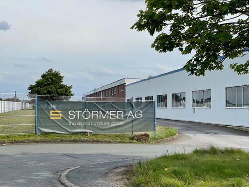 Einfahrt zum Firmengelände der Störmer AG mit großem Banner am Zaun vor einer modernen weißen Industriehalle an einer ruhigen Straße