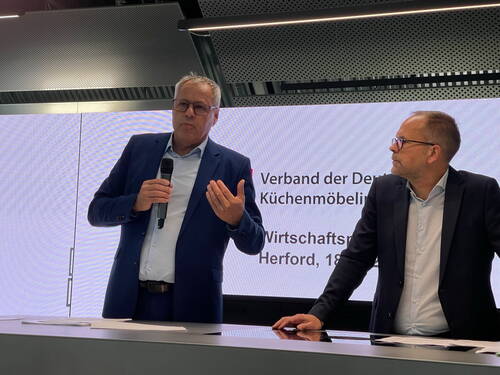 Zwei Männer im Anzug stehen an einem Rednerpult, während der linke mit Mikrofon in der Hand vor einer Leinwand mit der Aufschrift „Verband der Deutschen Küchenmöbelindustrie“ spricht und der rechte ihm aufmerksam zuhört.