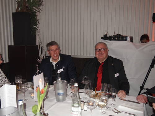 Zwei ältere Männer in Anzug und roter Krawatte sitzen lächelnd an einem festlich gedeckten Restauranttisch mit Kerzen, Tulpen und Weingläsern.