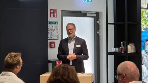Älterer Mann im Anzug hält einen Vortrag vor einer kleinen Zuhörergruppe in einem modernen, hellen Konferenzraum.