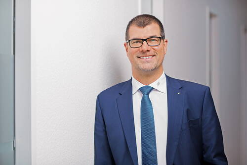 Lächelnder Geschäftsmann mit Brille im blauen Anzug lehnt an einer weißen Wand in einem modernen Büroflur
