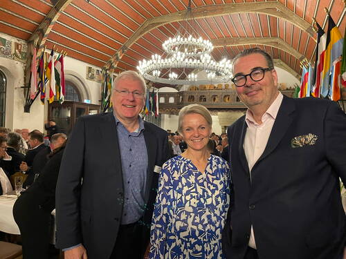 Drei elegant gekleidete Personen lächeln in einem festlich geschmückten Saal mit historischen Deckenmalereien, Kronleuchtern und zahlreichen Nationalflaggen im Hintergrund.