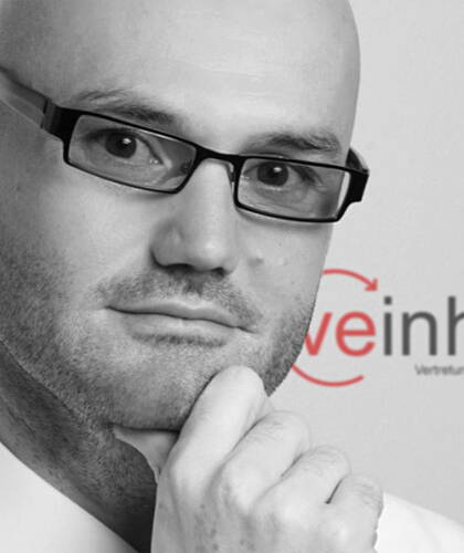 Nachdenklicher glatzköpfiger Mann mit Brille stützt sein Kinn mit der Hand vor einem unscharfen Firmenlogo im Hintergrund